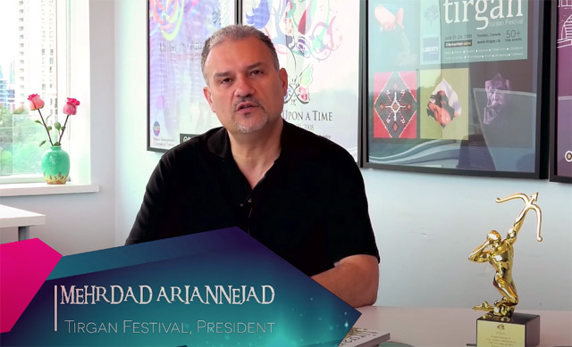 Mehrdad Ariannejad, as seen in a promotional video for the Tirgan Festival. He's sitting in an office with past festival posted on the wall an a statuette of a golden archer on his desk.