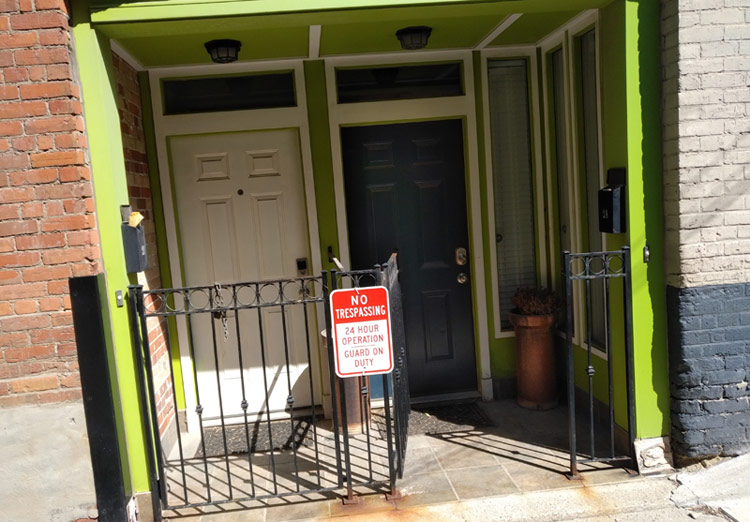 Two doors in an alcove framed by a neon green lintel. In front is a low-rising gate, to which is affixed a sign: "NO TRESPASSING – 24-Hour Operation – Guard on Duty"